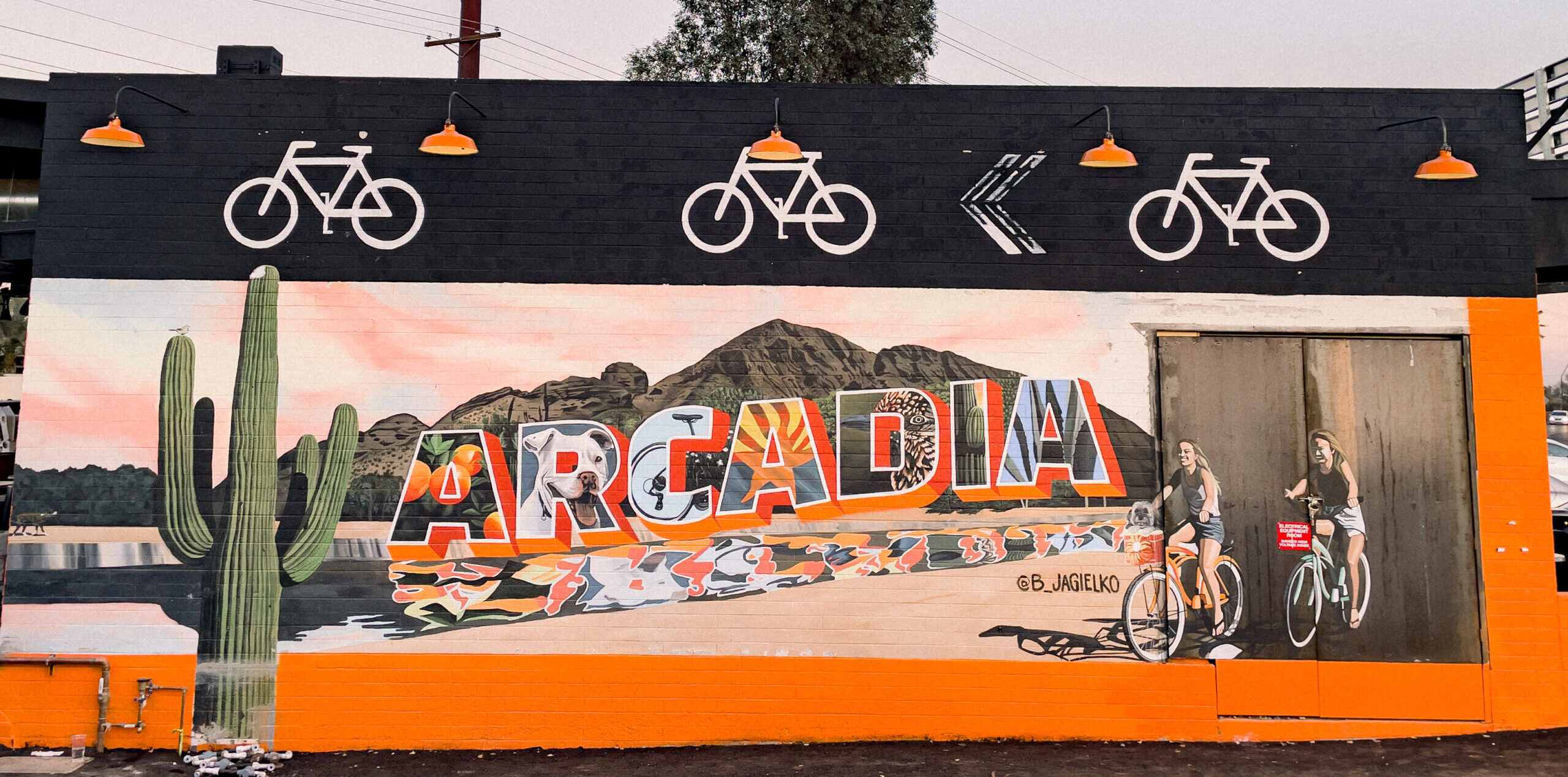 Hand painted mural of bicyclists and a desert landscape. The word Arcadia is in the center of the mural with Arizona inspired images in the center of each letter.